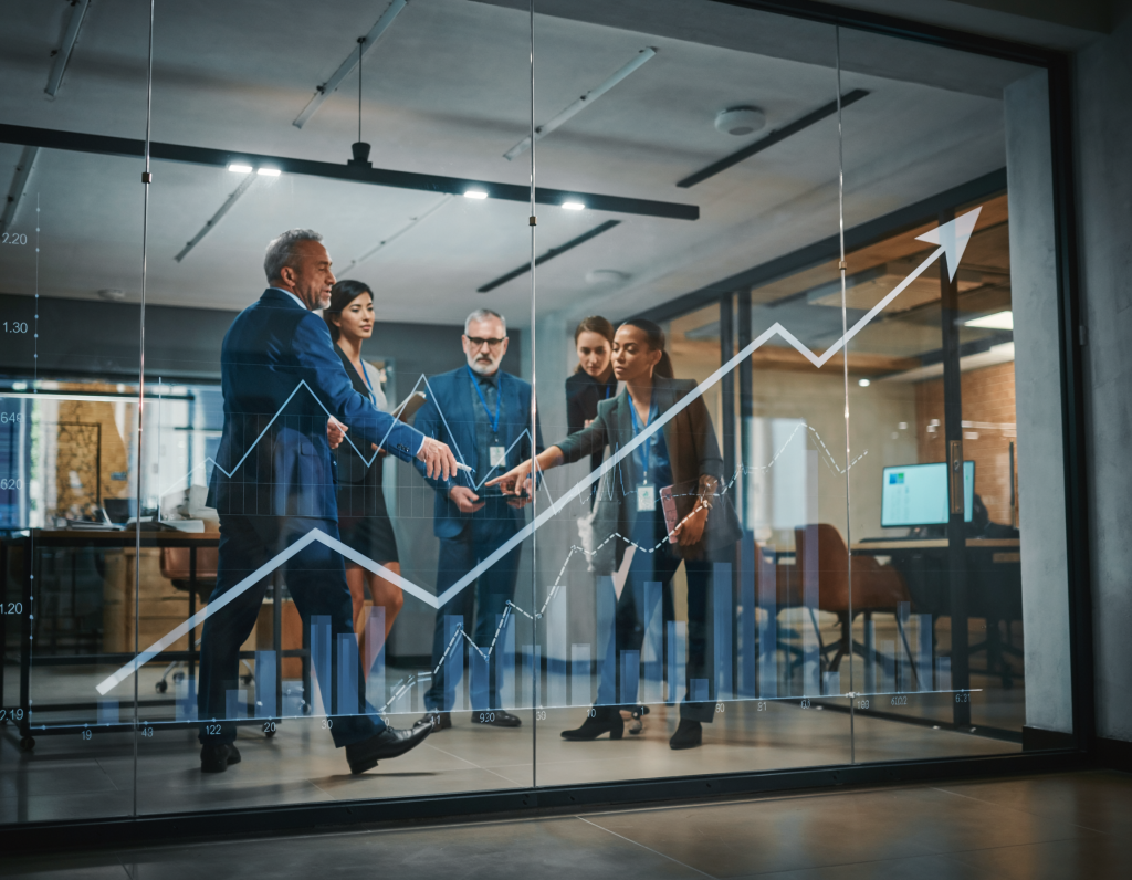 A realistic photograph of a business strategy meeting inside a modern UK manufacturing facility, with executives discussing data and AI performance metrics on a tablet or screen.