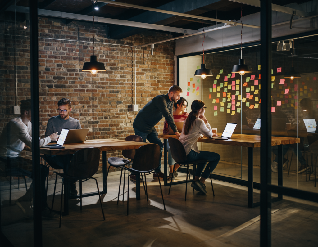 Cozy startup coworking space, exposed brick walls, wooden tables, MacBooks, sticky notes on glass boards, young team brainstorming, warm pendant lights