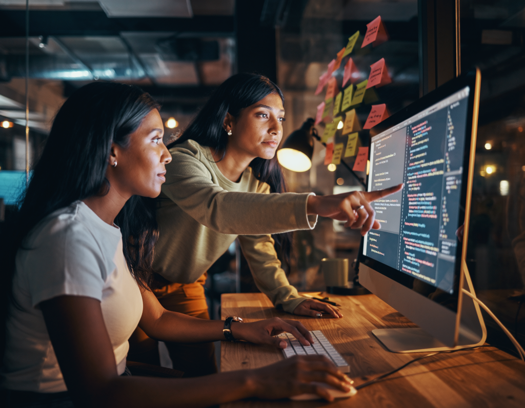 Full-stack developer pair programming, one typing, one pointing at screen, large iMac, sticky notes with user stories, casual startup vibe