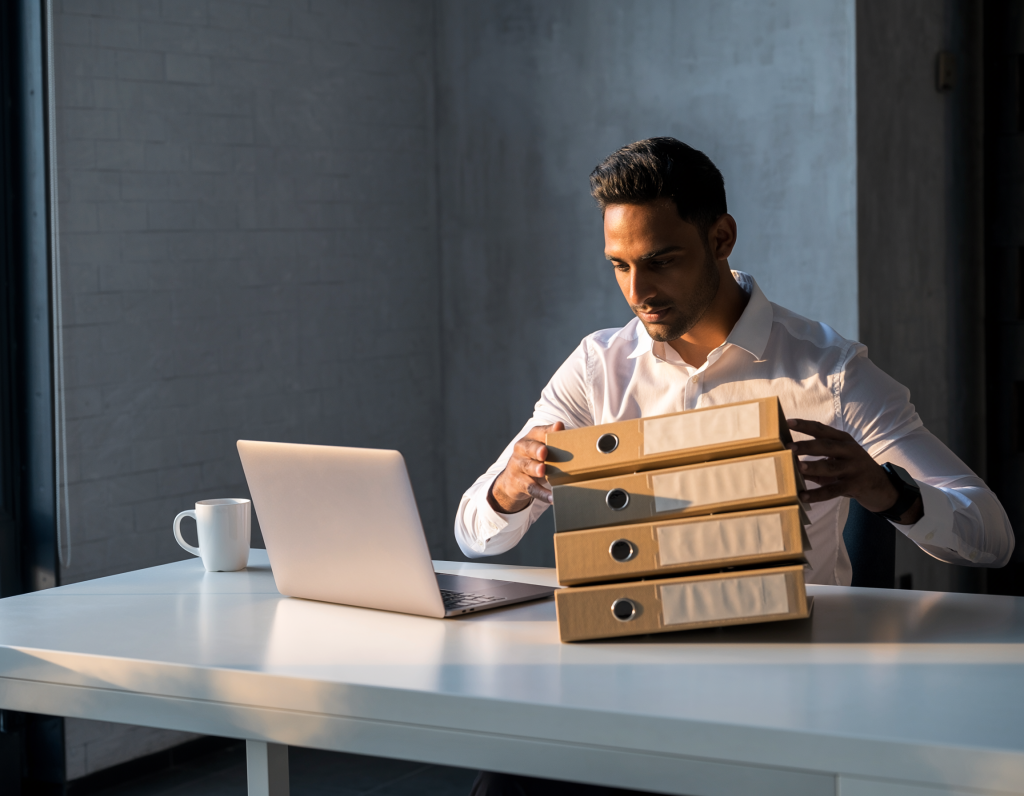 modern UK office, man seated at clean white desk calmly stacking three identical folders, closed silver laptop, white coffee mug, soft natural light from window, grey wall, professional photography, no text no logos no screen content, warm and orderly --ar 16:9